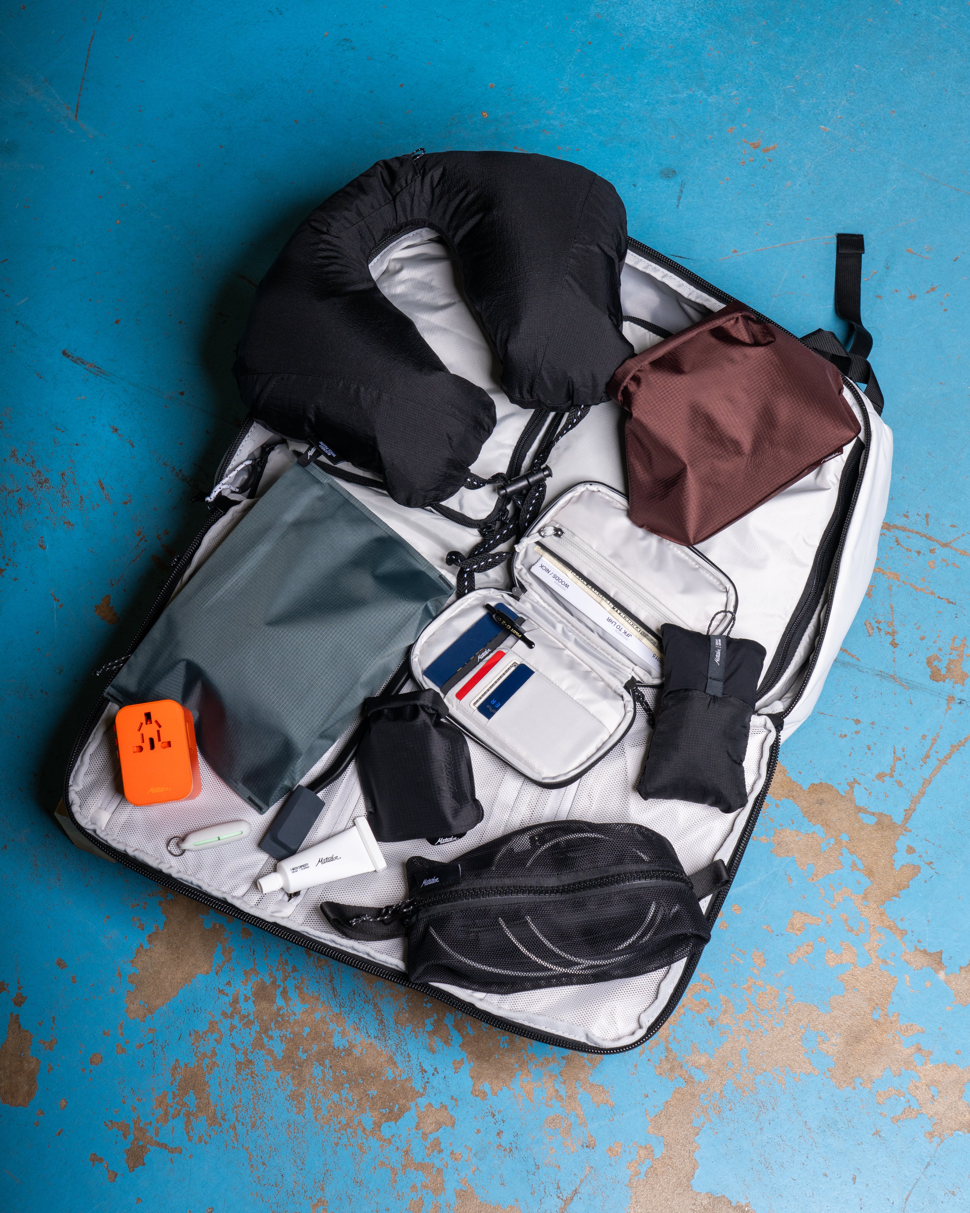 A white backpack being packed as luggage for international travel that has a collection of travel gear including a red toiletry case, global travel adapter, travel neck pillow, travel wallet, sleep mask, and travel-sized toiletries on a blue surface