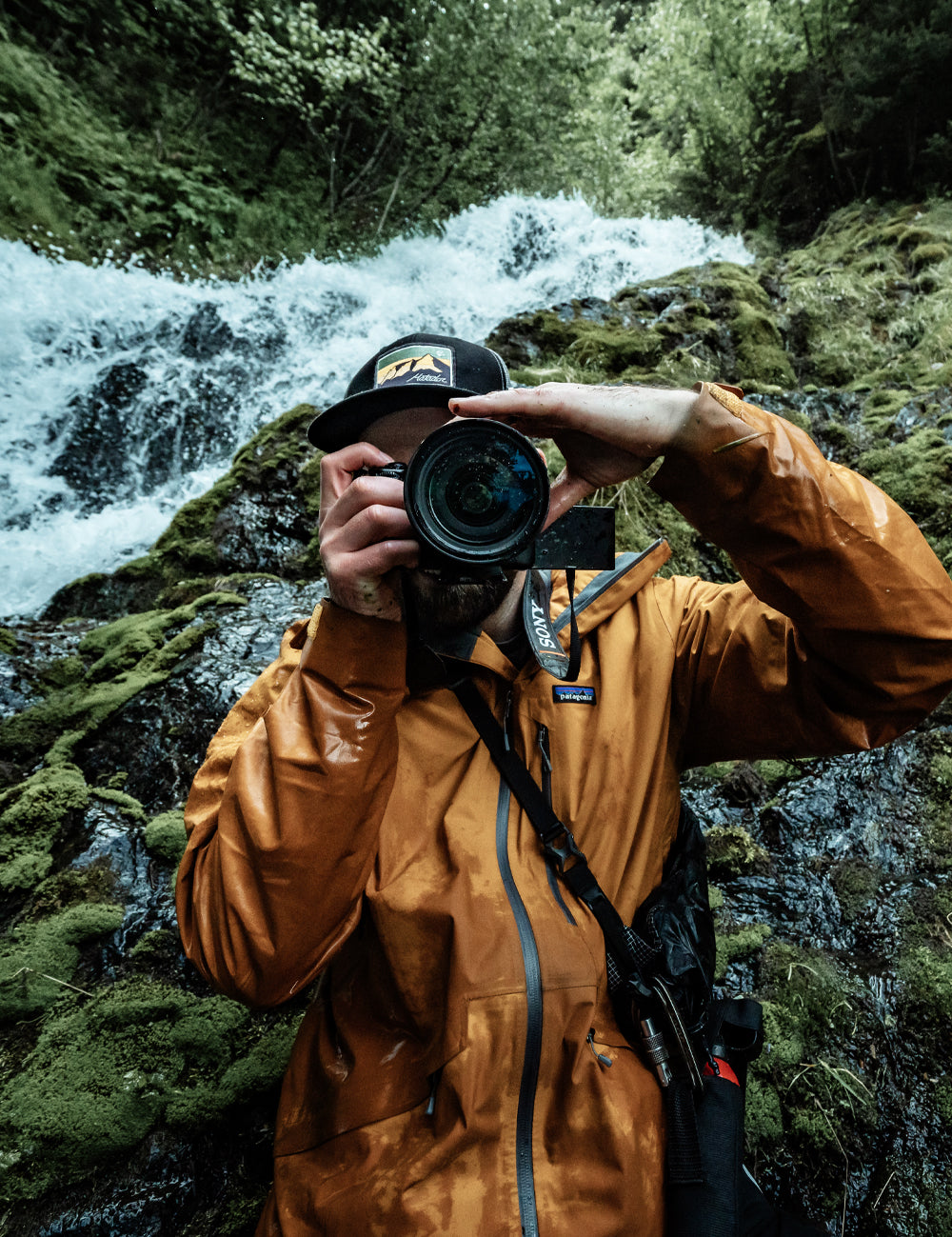 man in front of damp green waterfall looking through camera lens  