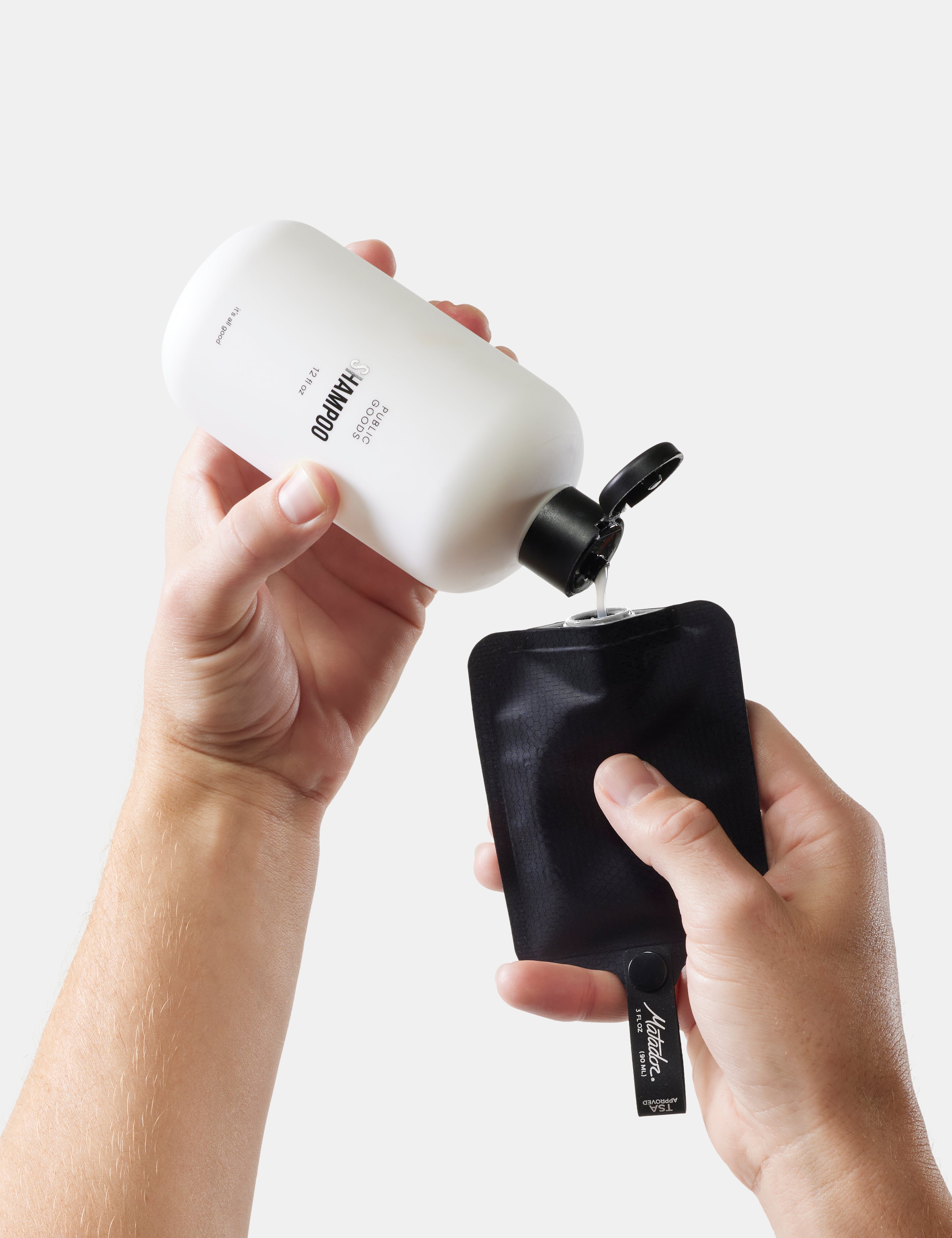 Person pouring liquid shampoo from a white bottle into a black travel-size toiletry bottle on a light gray background.