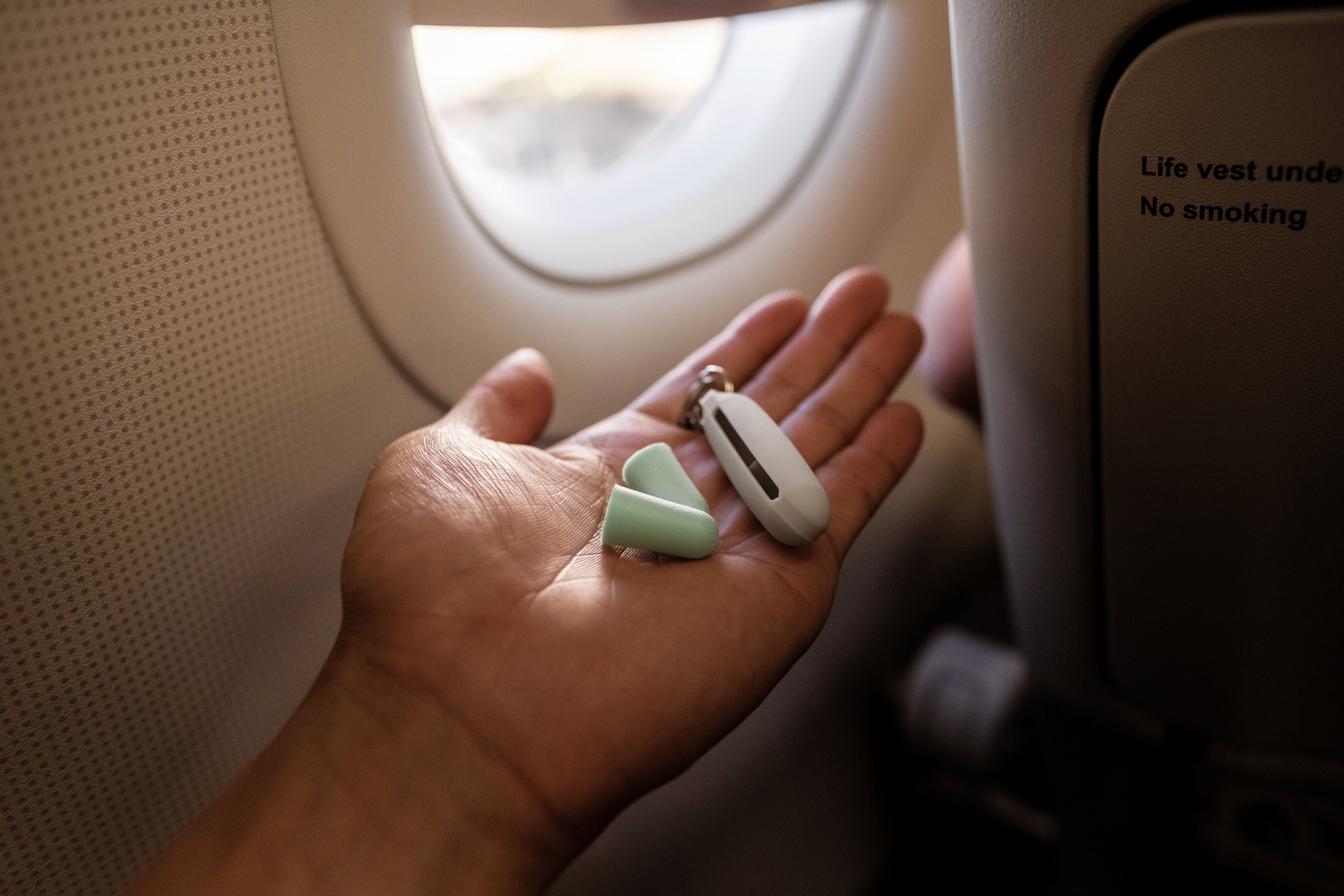 Hand holding two reusable ear plugs and a case with a keychain on an airplane in the window seat