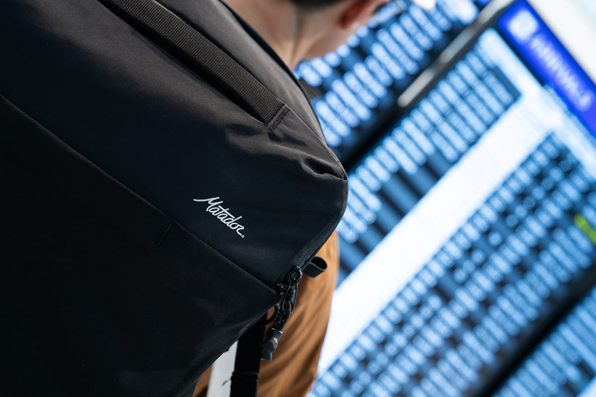 Person wearing a black backpack with Matador branding, standing in front of airport monitors preparing for an international trip.